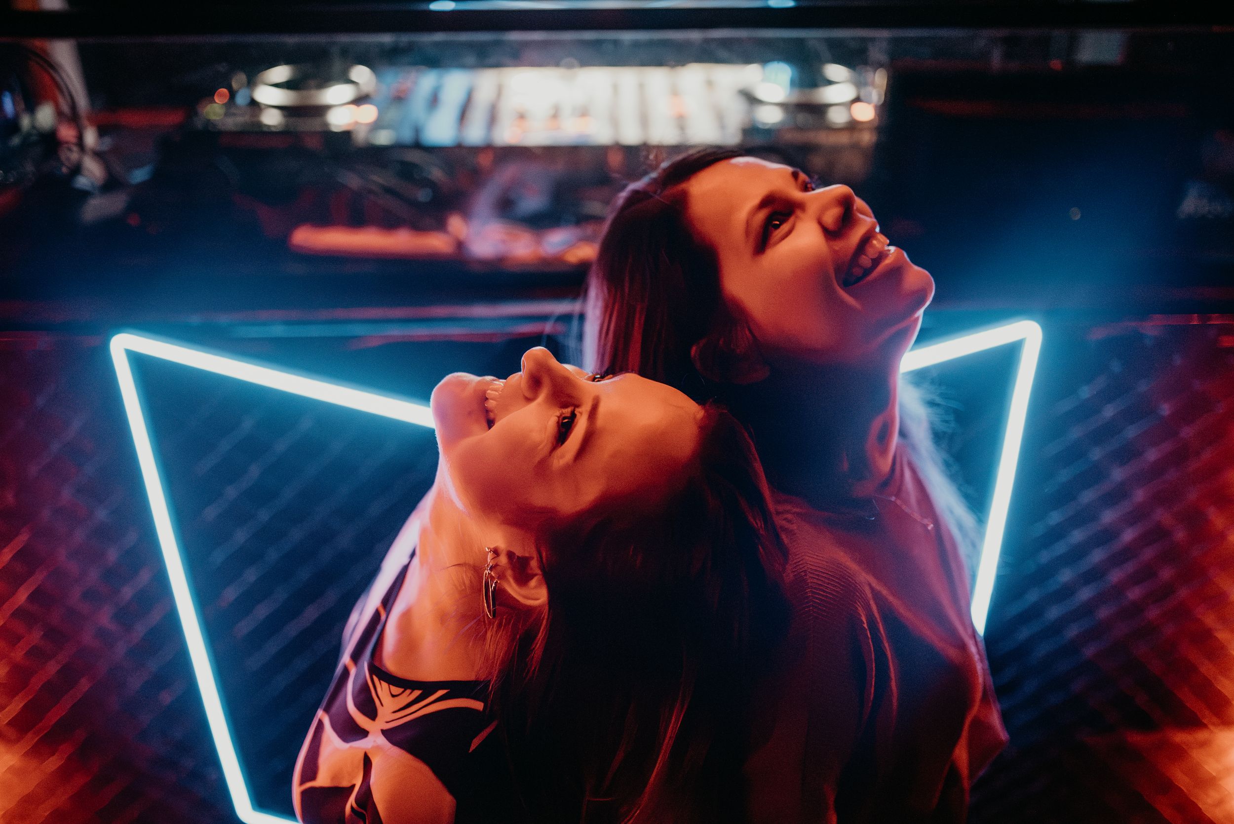 Two women laughing back to back in front of neon triangle light at nightclub. Fotógrafo de Bodas en Formosa