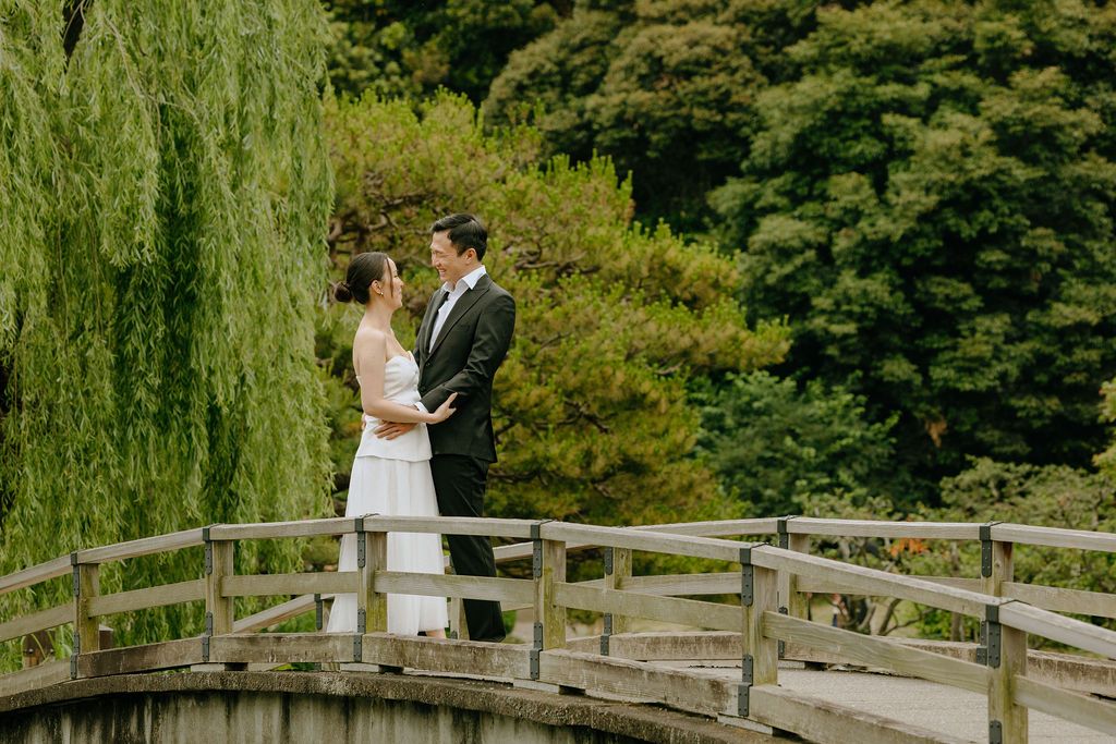 A bride and groom share an intimate moment on a wooden footbridge in a lush garden setting.