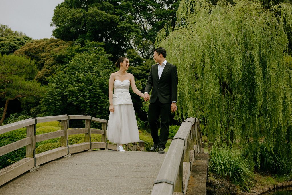 A couple stands on a wooden bridge in a serene Japanese garden with drooping willow trees.