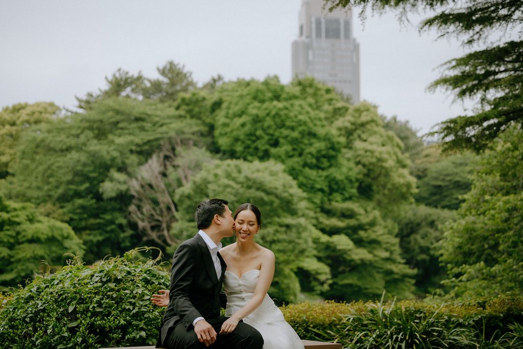 A couple in wedding attire sits together among green trees with a tower visible in the background.