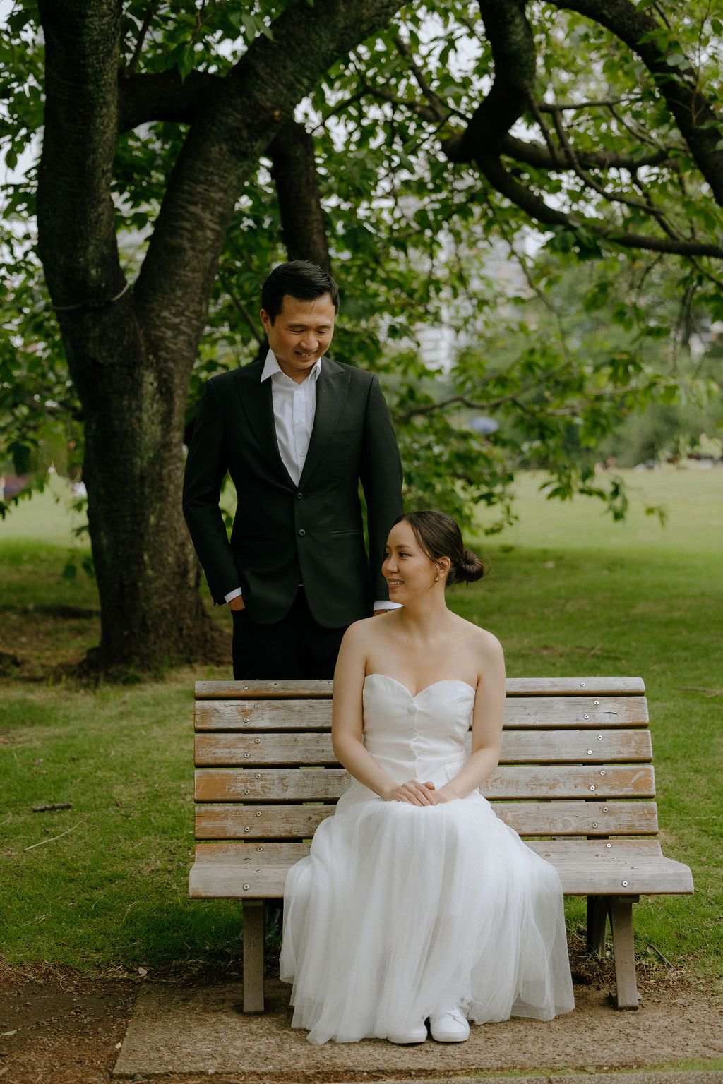 A couple shares a tender moment on a wooden bench surrounded by trees in a park setting.