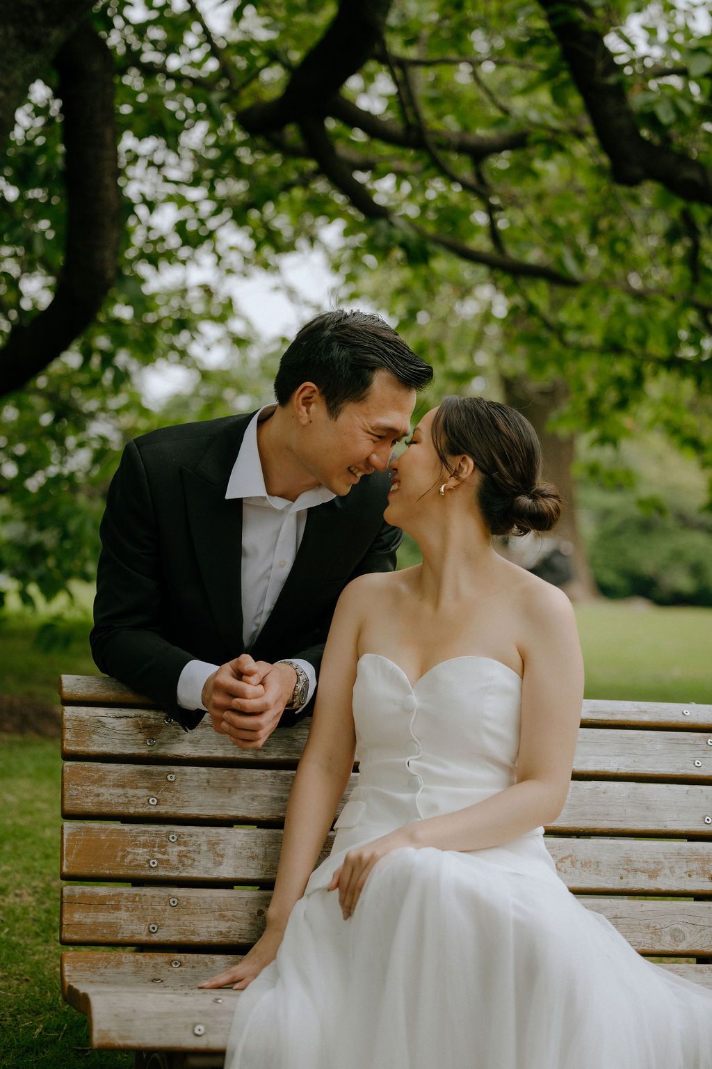 A romantic moment captured between newlyweds sitting on a park bench under green trees.