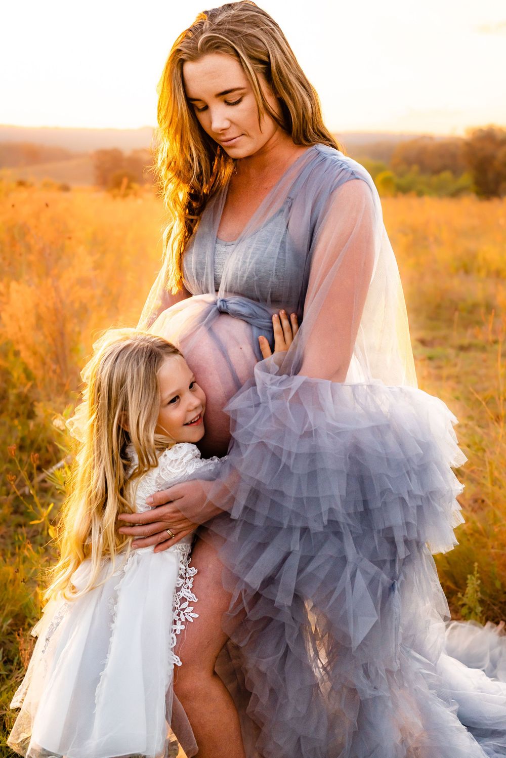 Maternity portrait with young girl embracing mother's belly in golden sunset light.