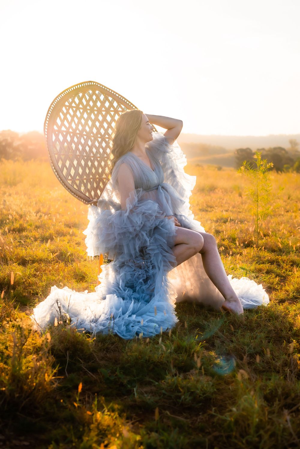 A peaceful maternity portrait on a peacock chair in a sunlit field with a flowing gray dress.