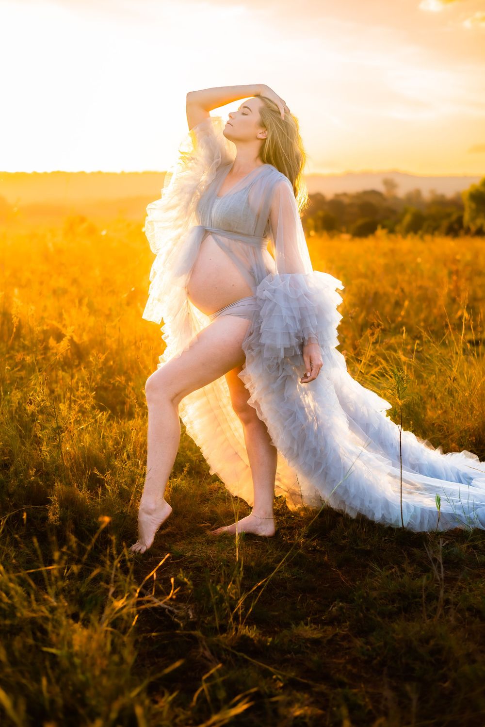 Dreamy maternity portrait with white ruffled gown in sunlit meadow.