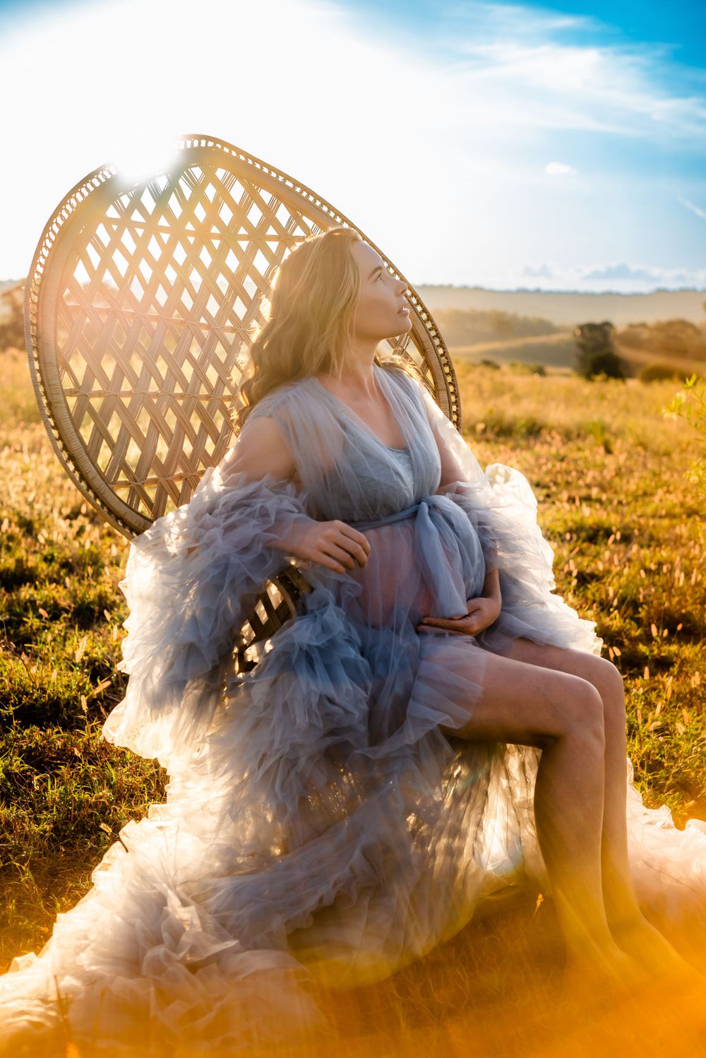 An ethereal maternity photo of a woman in a flowing gray dress seated on a peacock chair at golden hour.