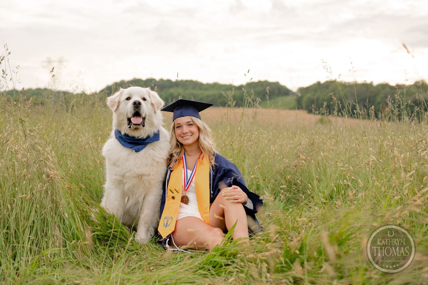 Graduate in cap and gown poses with their white dog wearing matching blue bandana in grassy field.