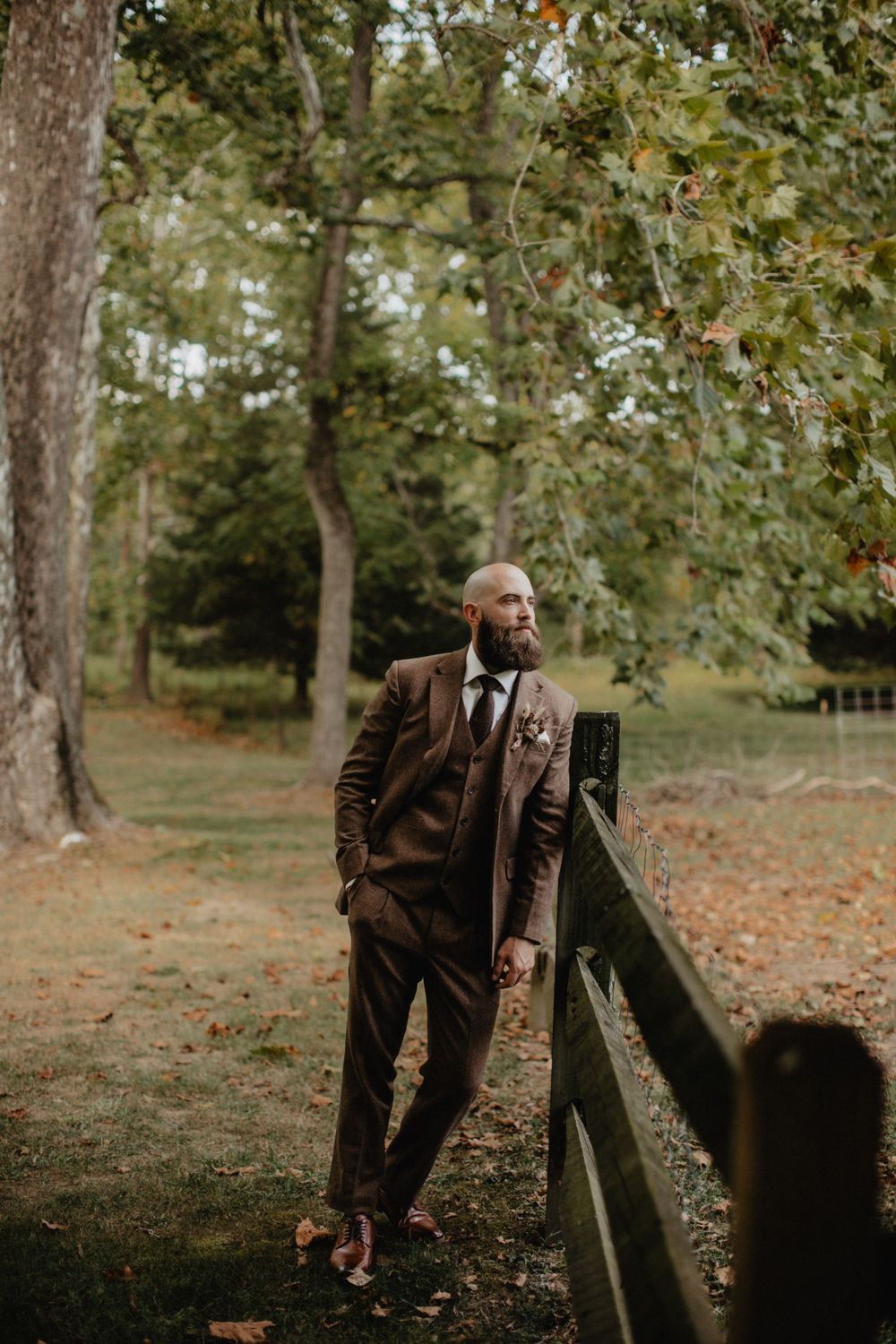 A person in a brown suit leans against a wooden fence in an autumn forest setting with fallen leaves on the ground. NJ Wedding Photographer
