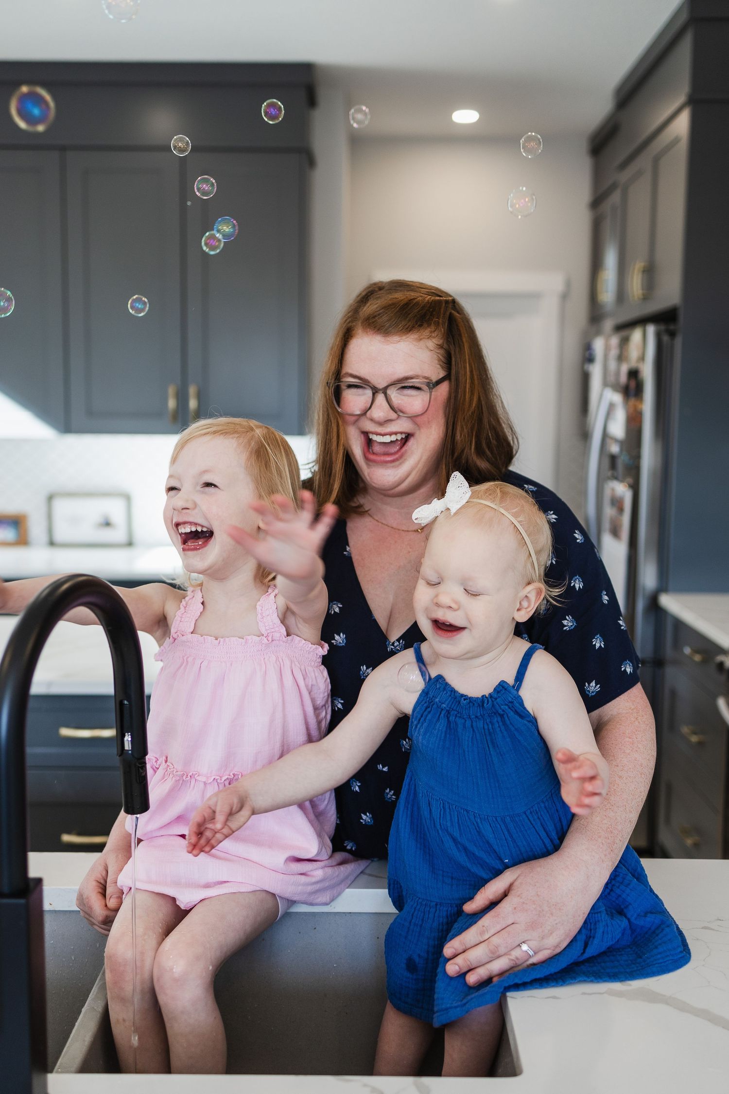 A mother and her two daighters delight in bubbles being blown toward them in the kitchen