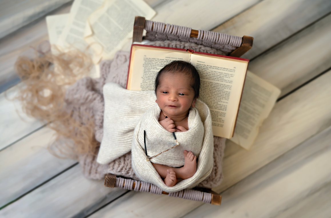 Newborn baby swaddled in white wrap, smiling while lying on an open book in a cozy photography prop setup. Embracing Life’s Beauty