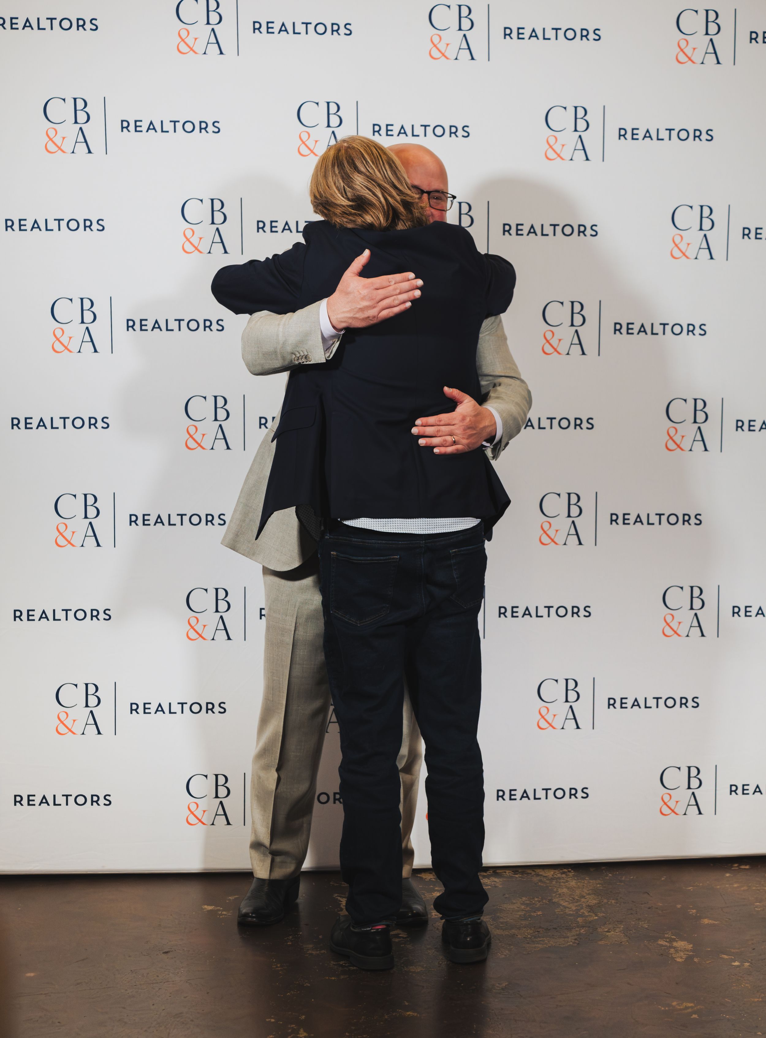 Two people share a warm hug in front of a CB&A Realtors branded backdrop at a professional event.