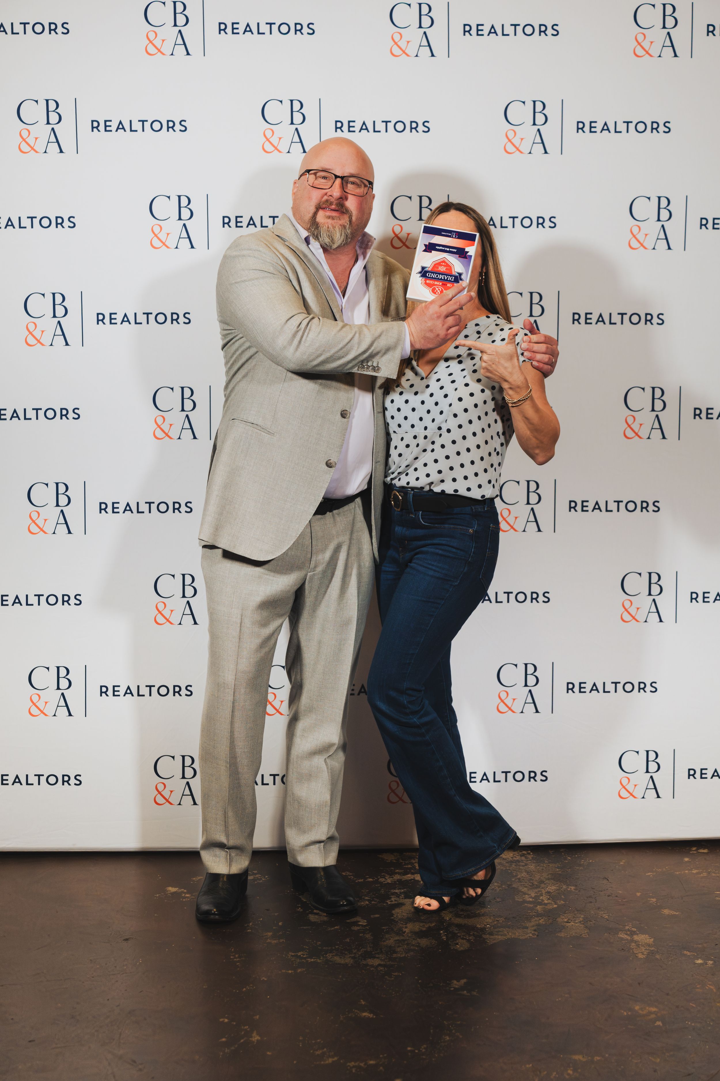 A man and woman pose playfully in front of a CB&A Realtors branded step-and-repeat backdrop at a real estate event.