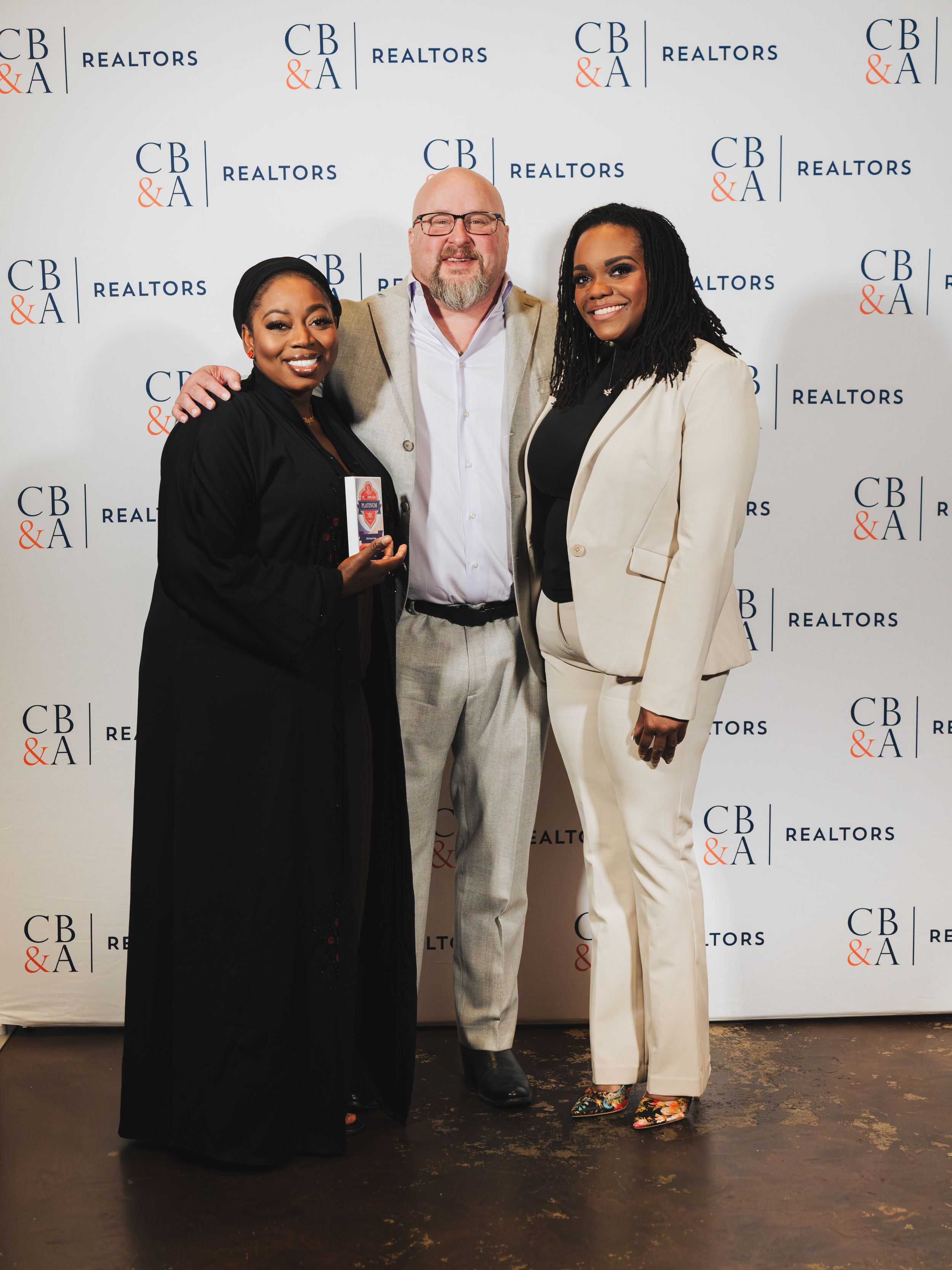 Three professionals pose together in front of a CB&A Realtors branded backdrop at an awards event.
