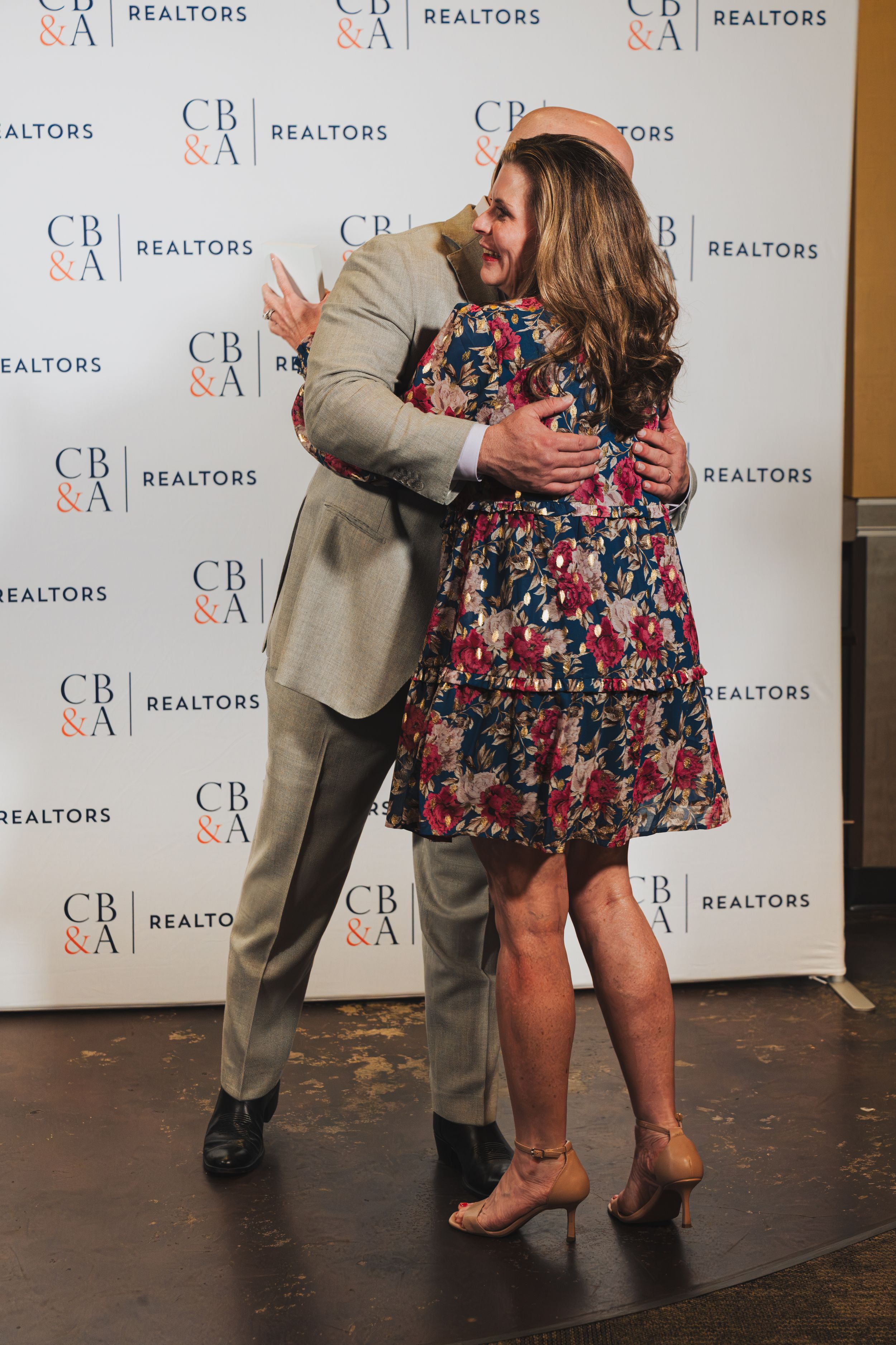 Two people share a warm hug at a CB&A Realtors event, one in a floral dress and the other in a tan suit.