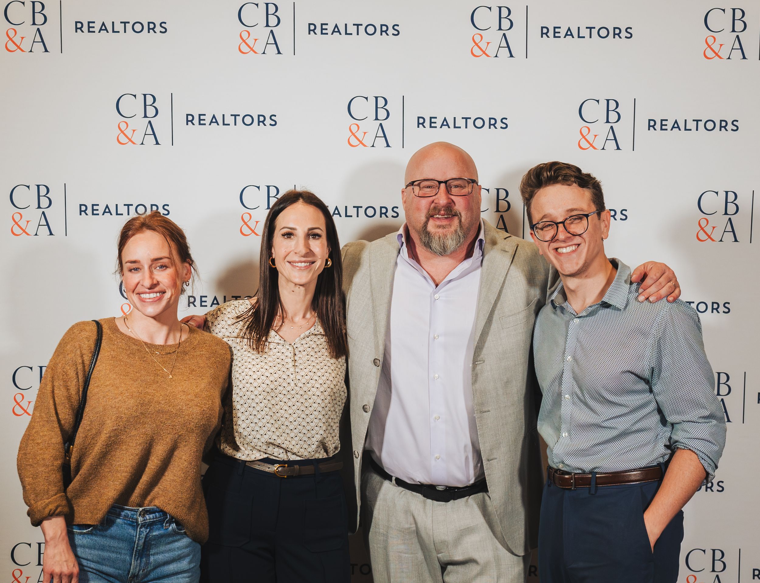 Four professionals smiling together at a CB&A Realtors corporate event backdrop.