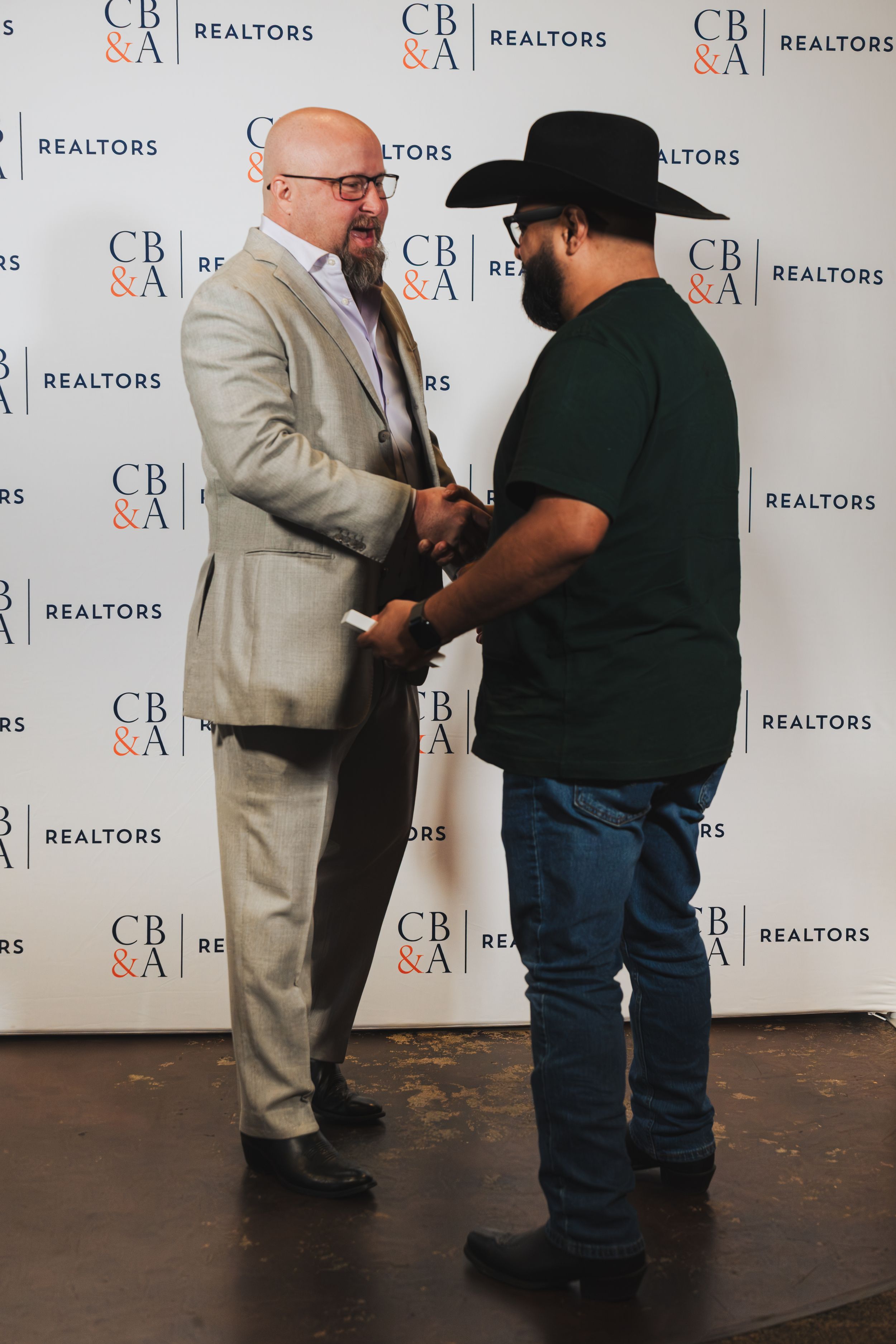 Two men shaking hands in front of a CB&A Realtors branded backdrop at a professional event.