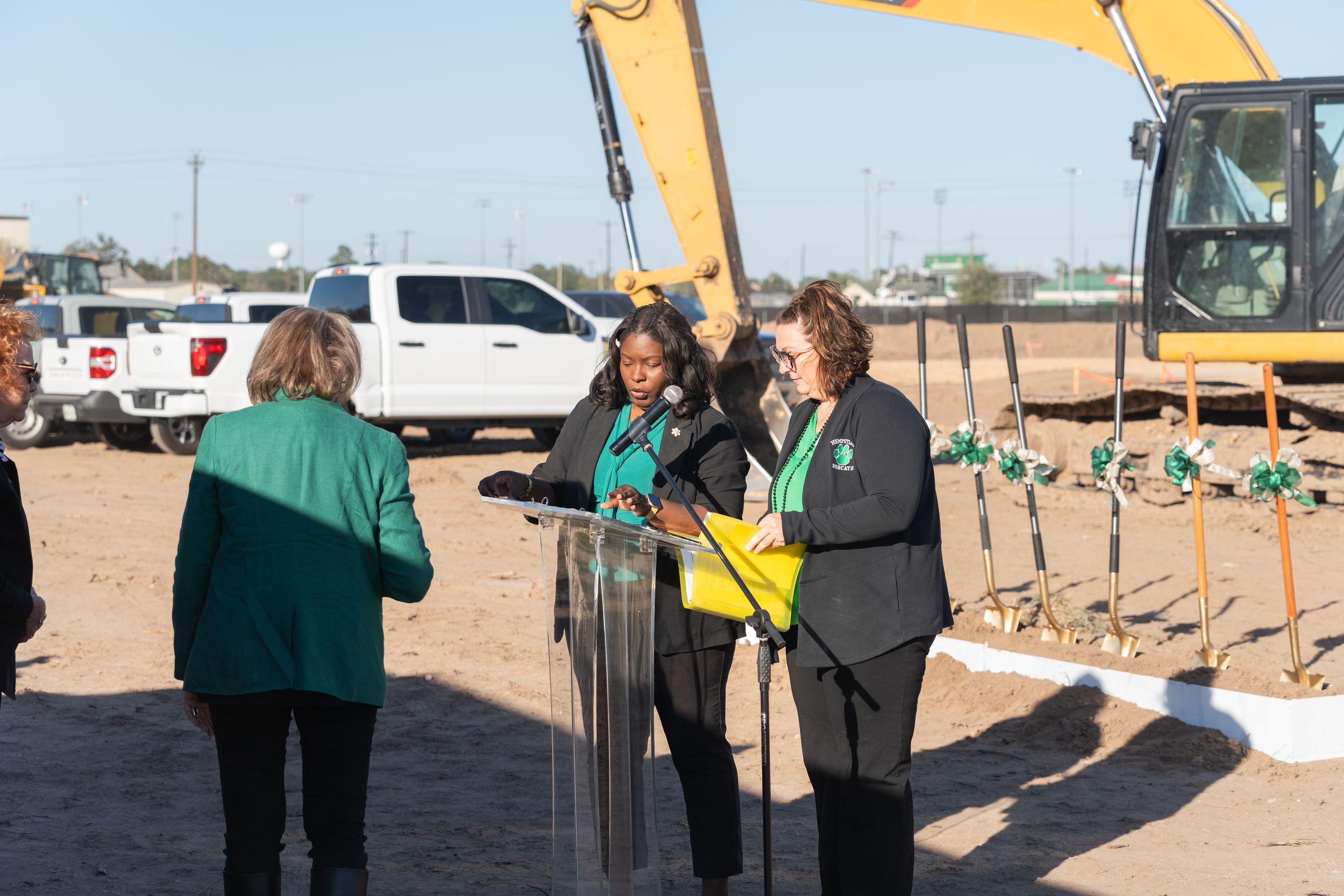 People gather at a construction site with excavator equipment and vehicles in the background.