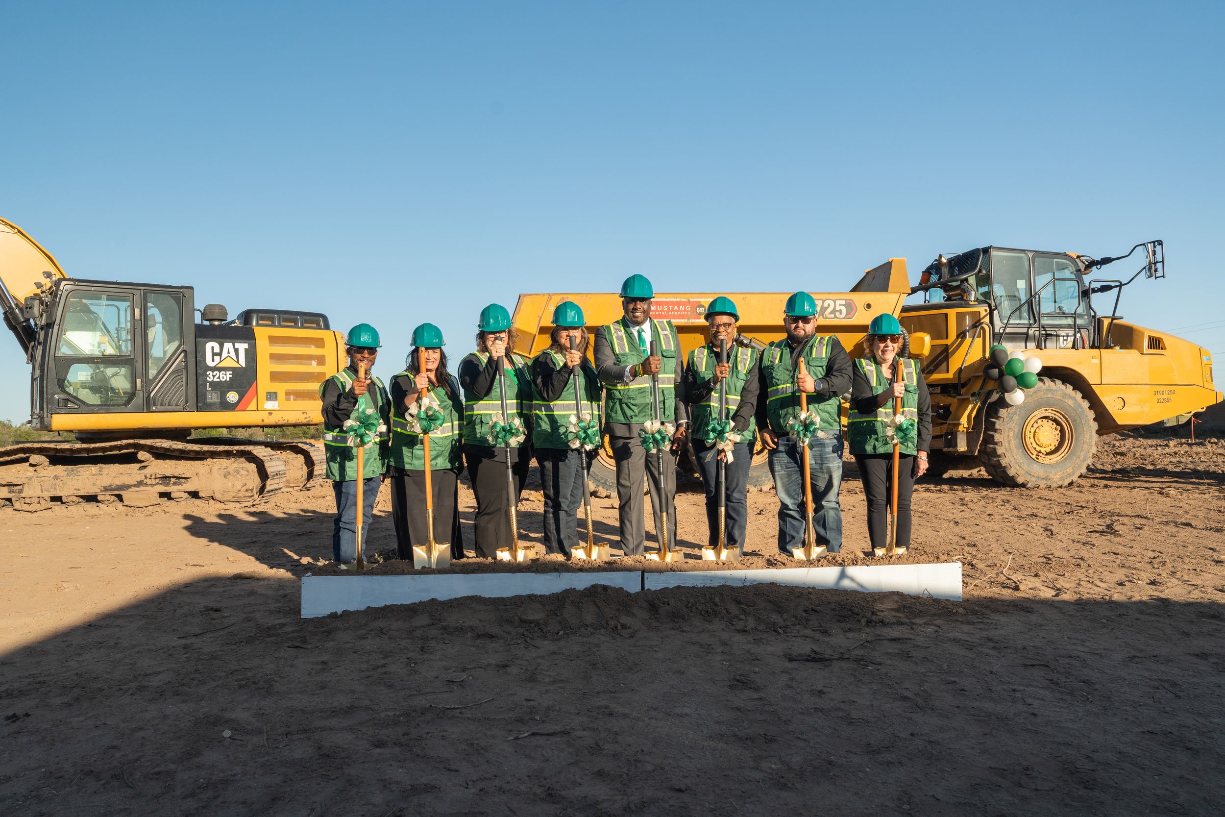 A line of workers in safety gear stand against a desert backdrop with heavy construction equipment behind them.