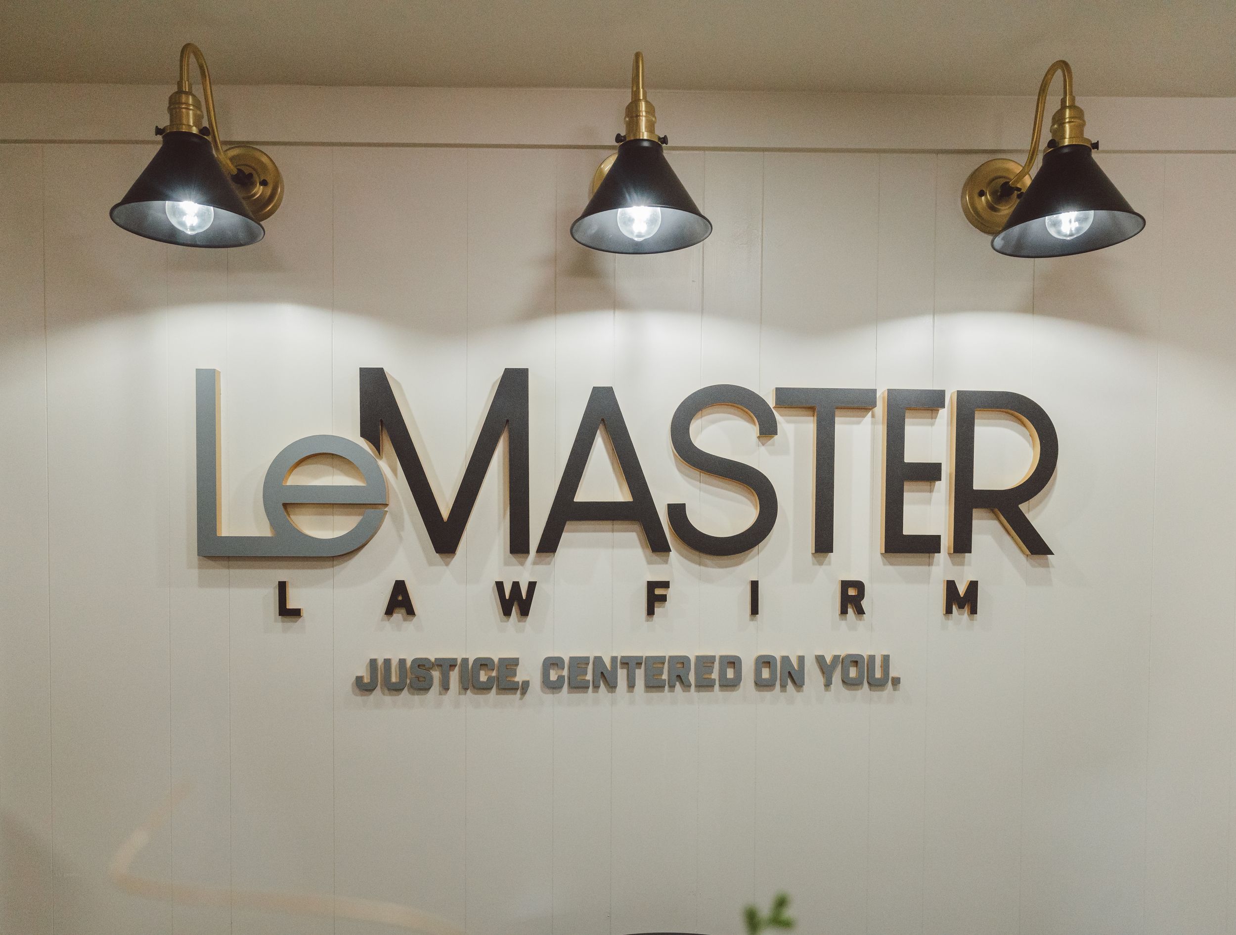 Law firm office sign with "LEMASTER" text and tagline "JUSTICE, CENTERED ON YOU" mounted on light colored wall.