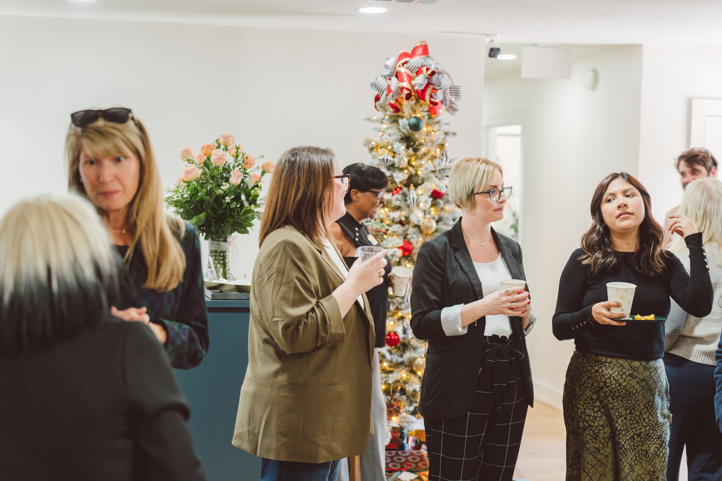 A group of people enjoying drinks and conversation at a holiday party with a decorated Christmas tree in the background.