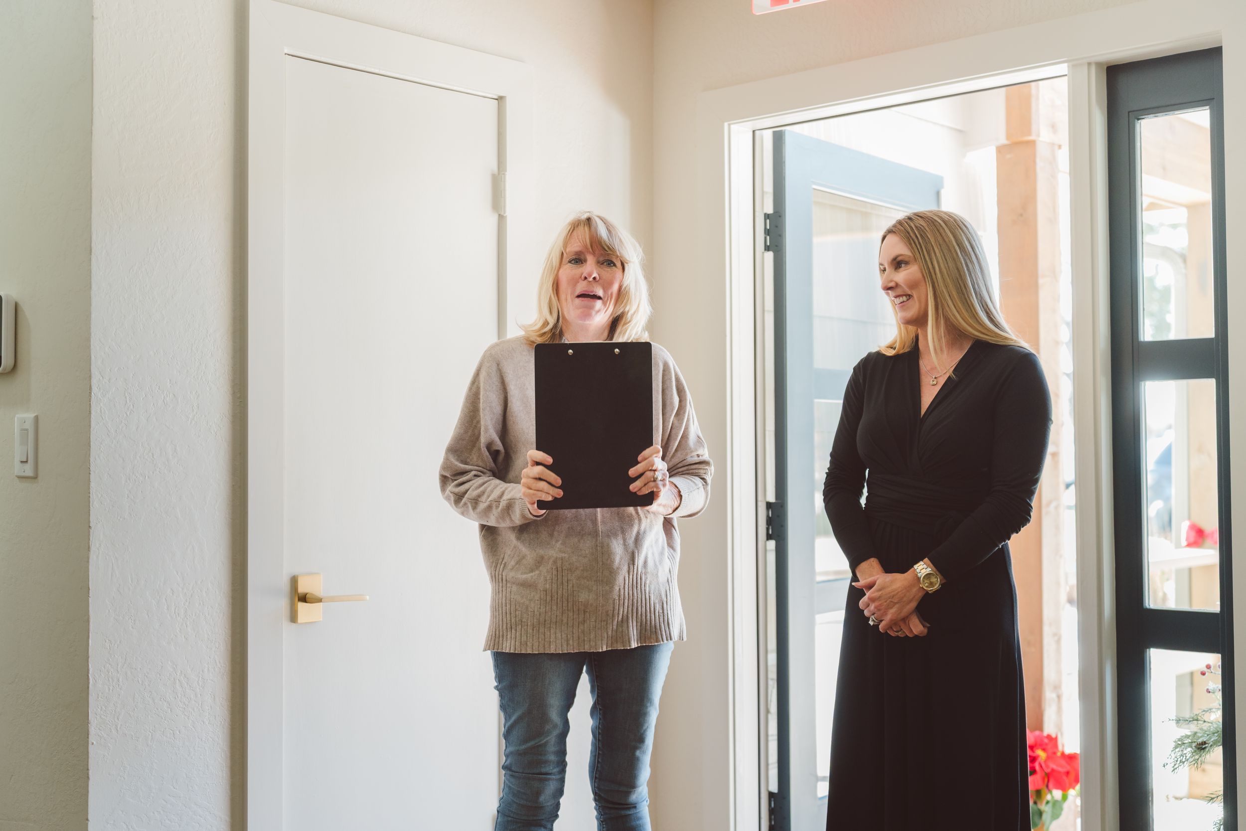 Two people stand in an office doorway, one holding a clipboard while wearing a beige sweater.