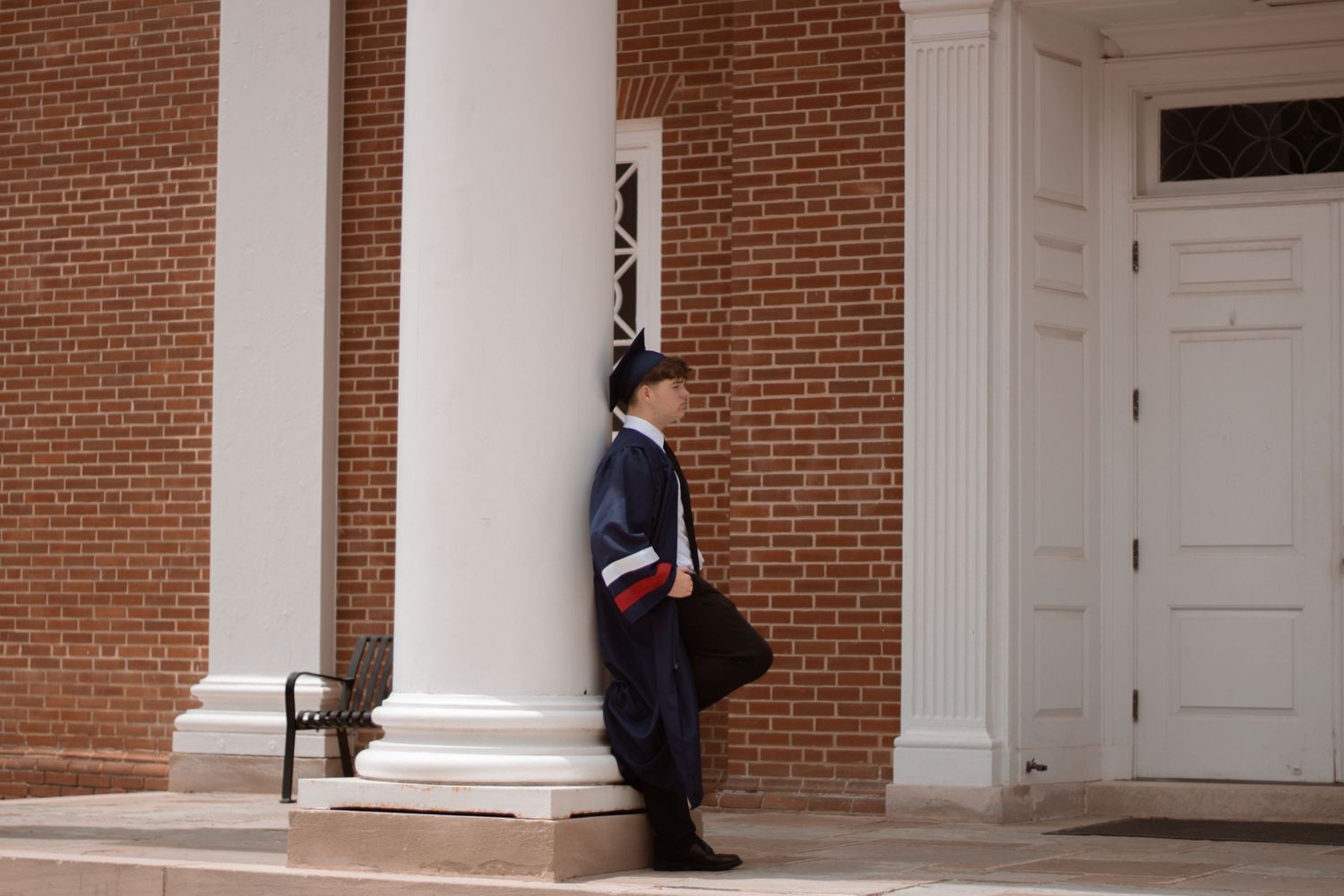 Graduate in academic regalia poses against a classic brick and white column building facade.
