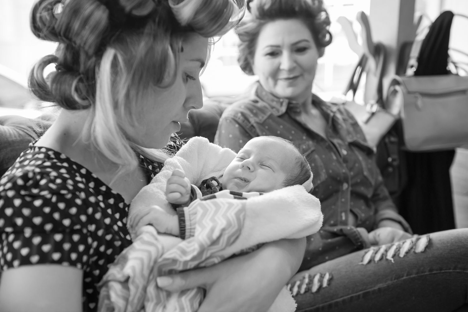 Women with vintage hairstyles in polka dot dresses hold and admire a newborn baby.