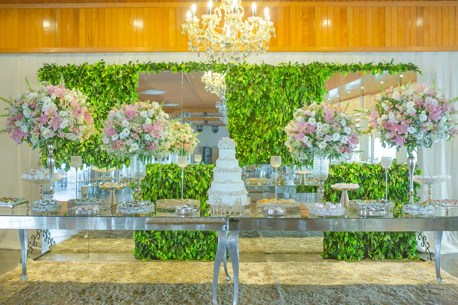 Elegant wedding dessert table display with white tiered cake, floral arrangements, and greenery wall backdrop.