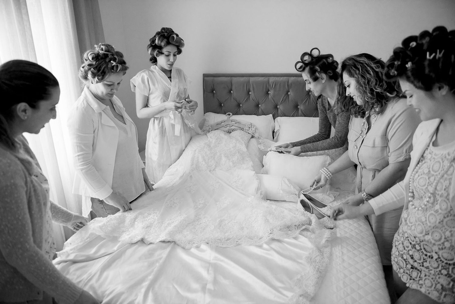 Black and white photo of wedding dress preparation with group gathered around bed.