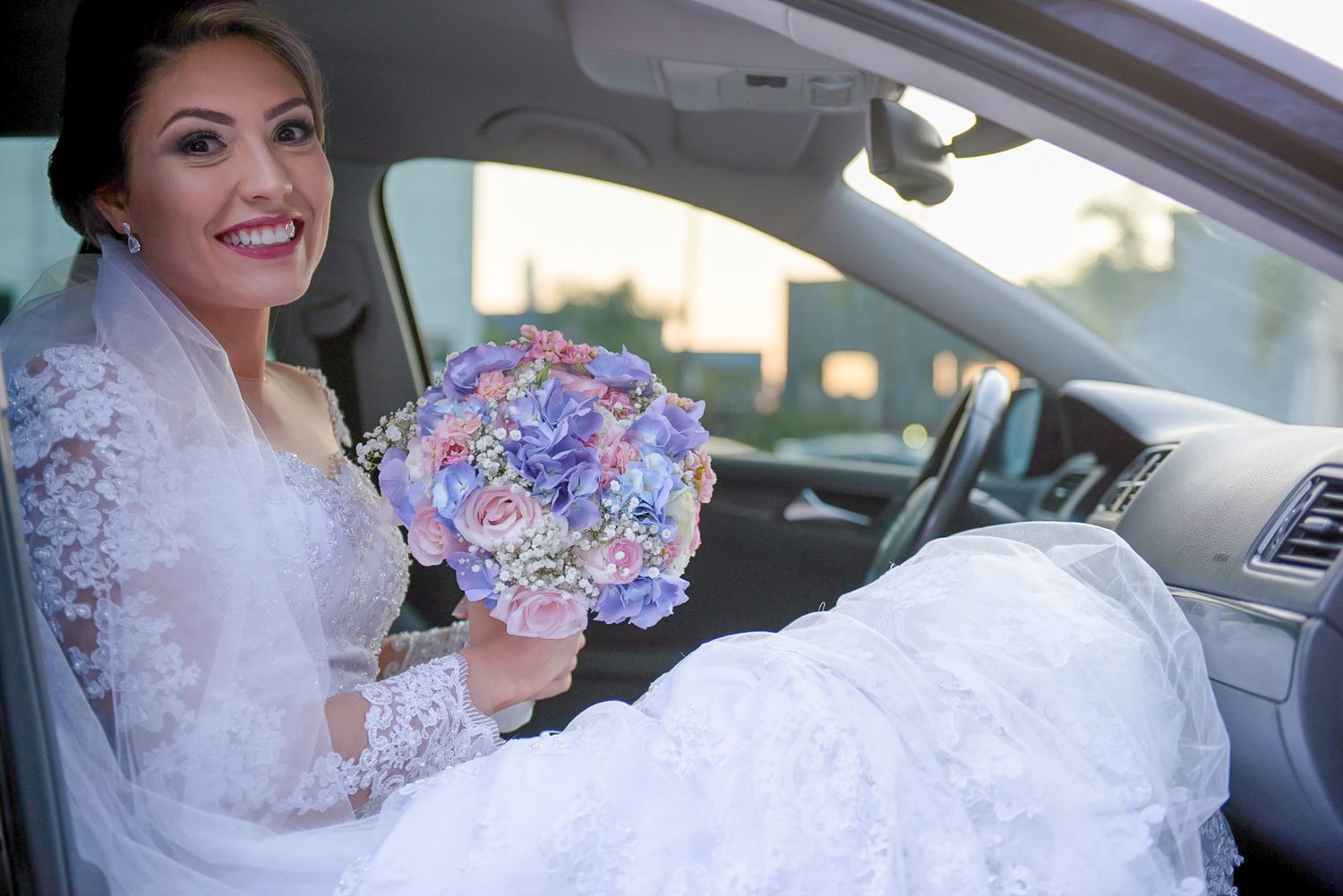 A joyful bride holding a purple and blue bouquet while seated in a car.