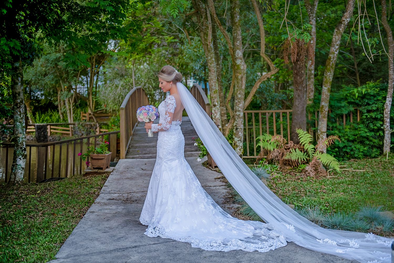 A wedding photo of a bride in a lace dress with long train walking on a wooden boardwalk surrounded by greenery.