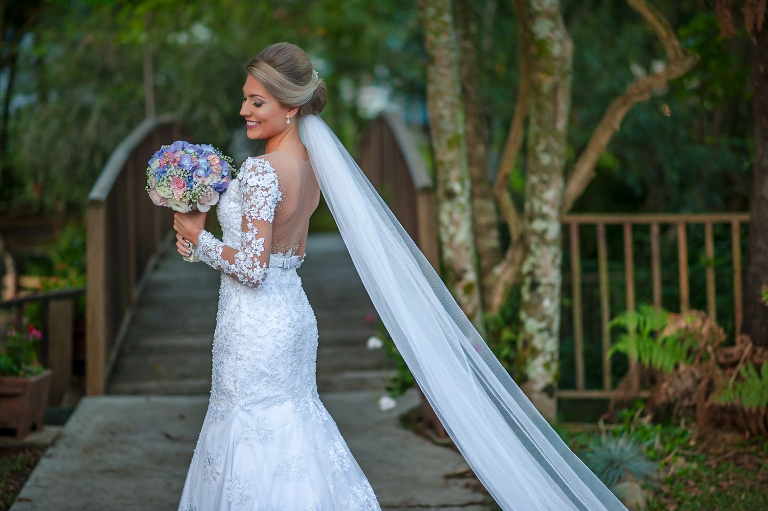 Bride in fitted lace gown holds purple bouquet while posing on wooden walkway in garden setting.