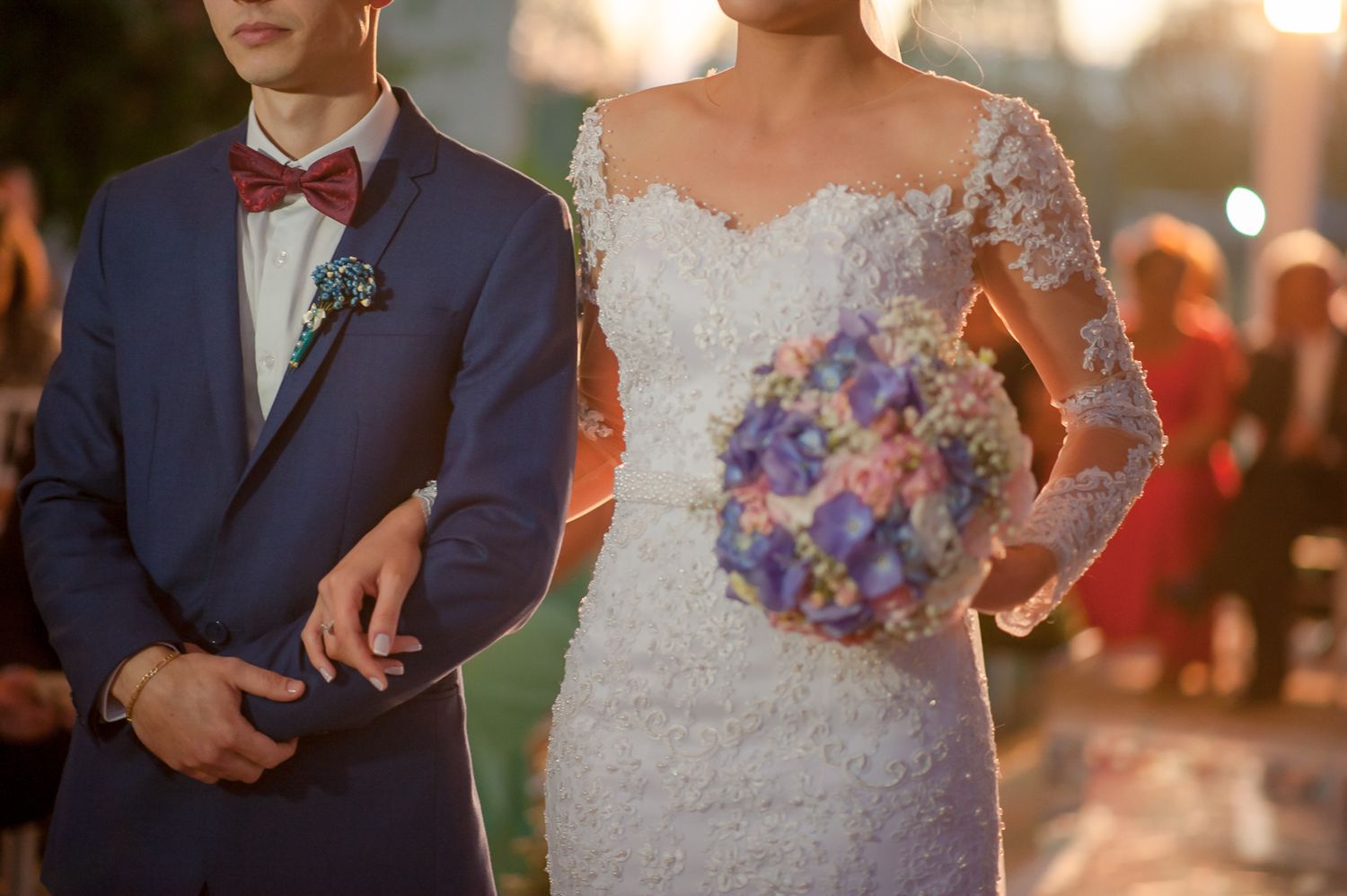 Close-up of wedding attire showing navy suit with bow tie and lace dress with purple bouquet.