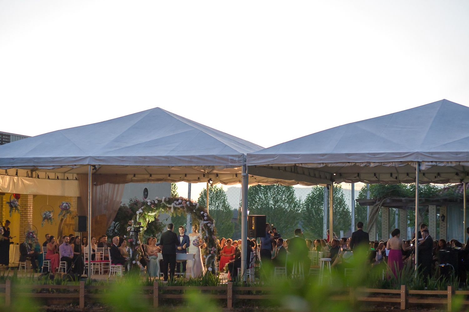 Outdoor wedding reception setup with white tents visible through trees at dusk.