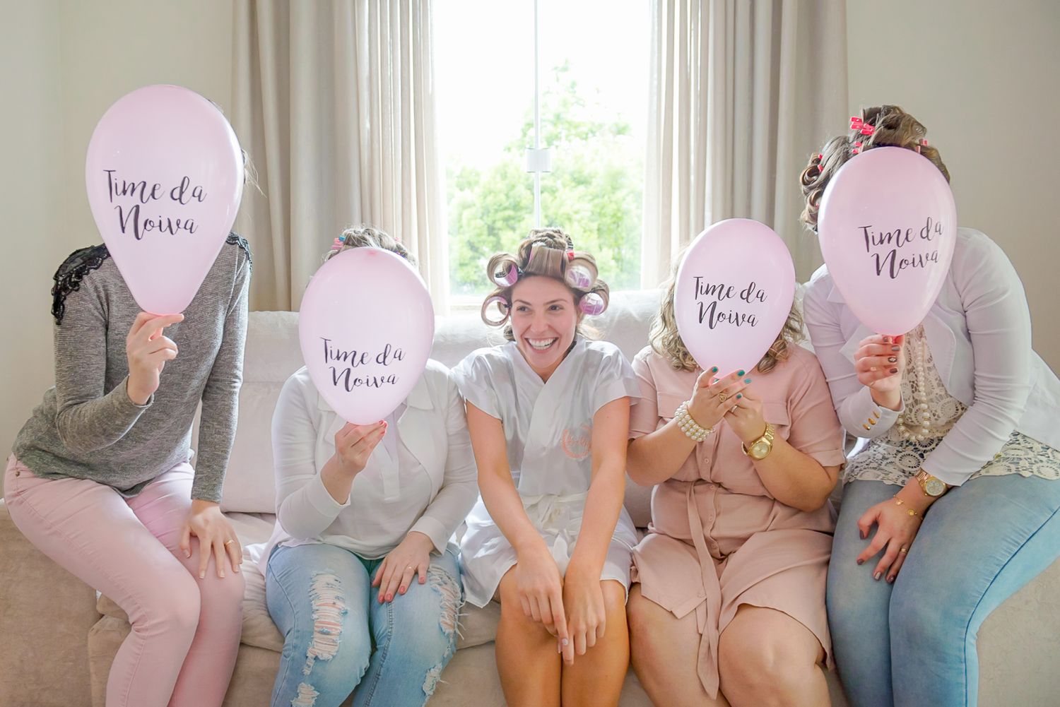 A group sits on a couch holding pink balloons that say "Here Comes Florence" at a bridal celebration.