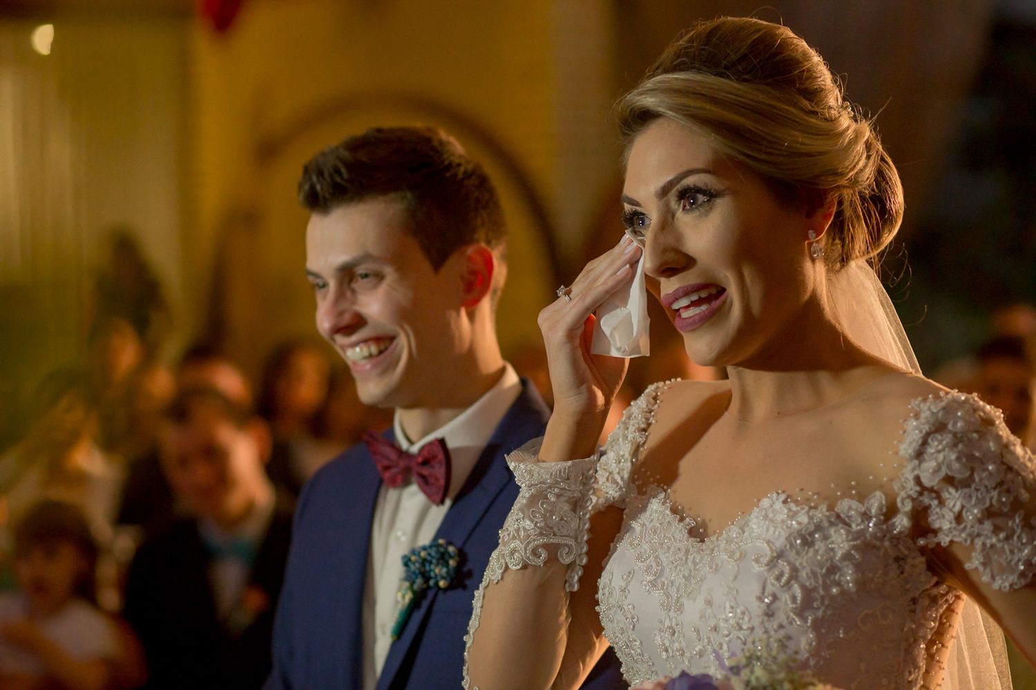 Emotional moment captured during wedding ceremony with warm lighting and crowd in background.