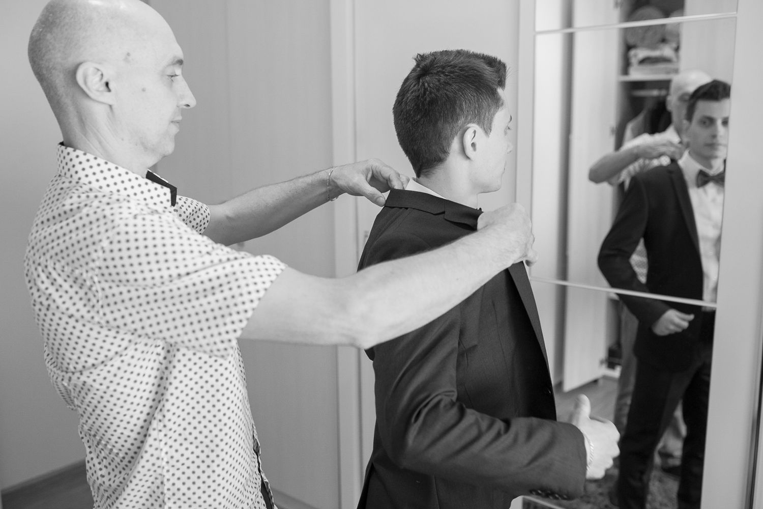 A stylist helps adjust the back of a groom's formal attire in front of a mirror.