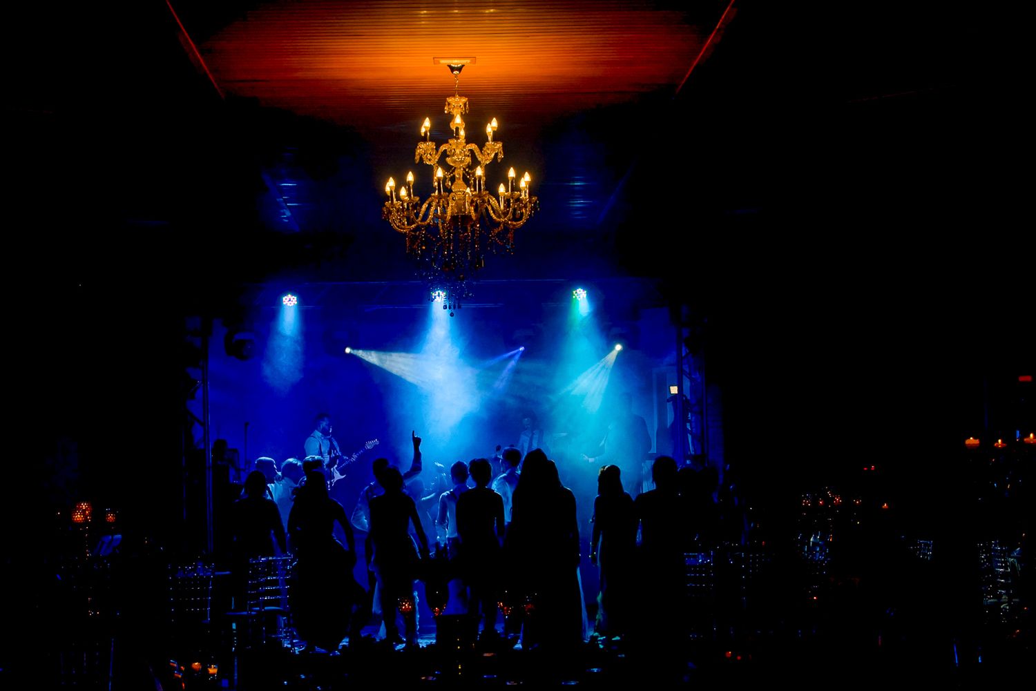 Wedding reception dance floor illuminated by blue spotlights and chandelier in dark venue.