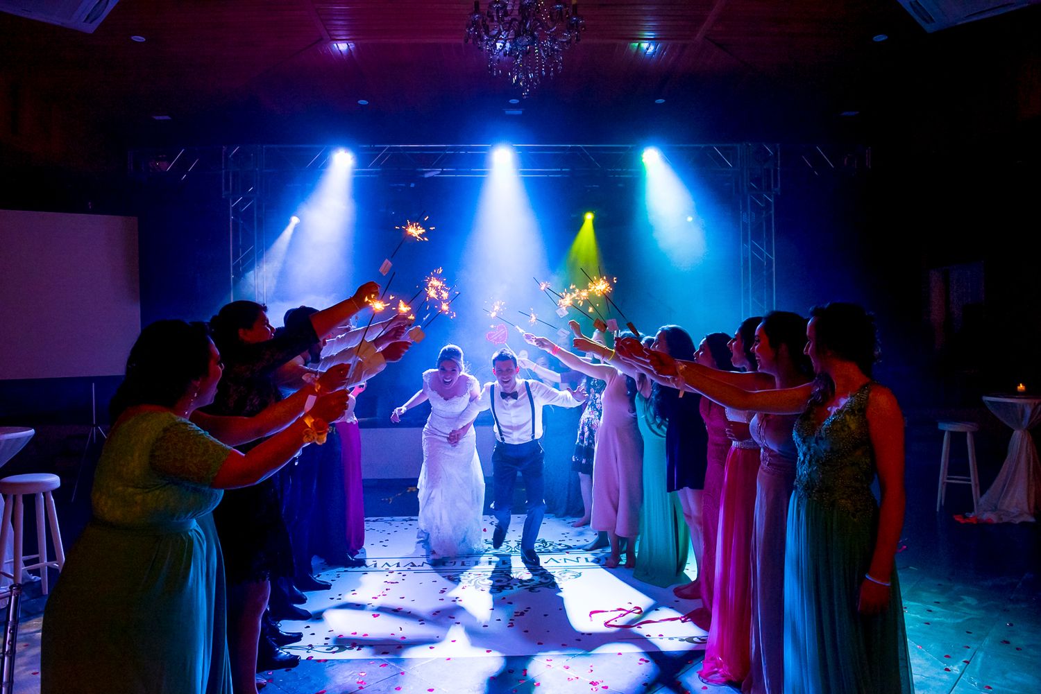 Wedding party forms sparkler tunnel for dancing couple in blue and purple lighting.