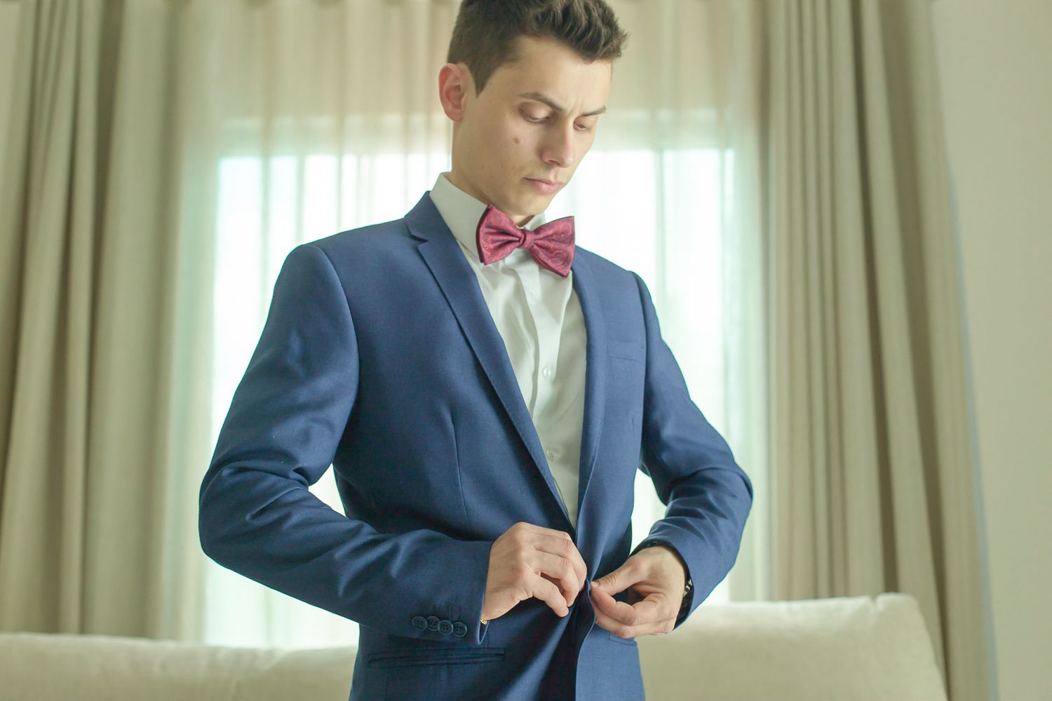 A groom in a navy blue suit and pink bow tie buttons his jacket while getting ready.