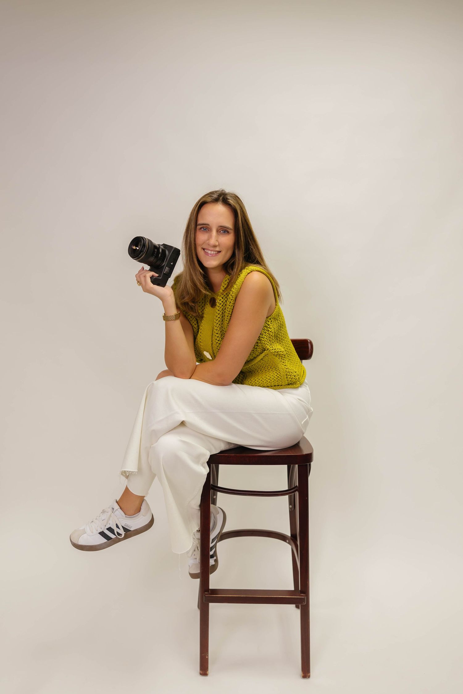 A photographer in bright yellow top and white pants poses with camera on wooden stool.