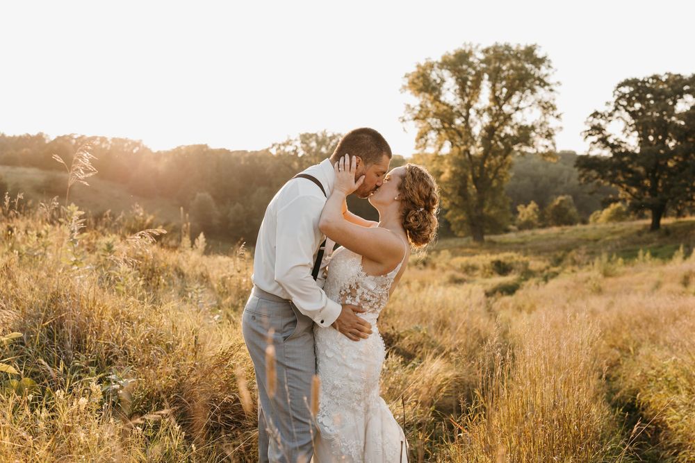Couple sharing a kiss in a sunlit golden meadow, groom in suspenders and bride in lace wedding gown.