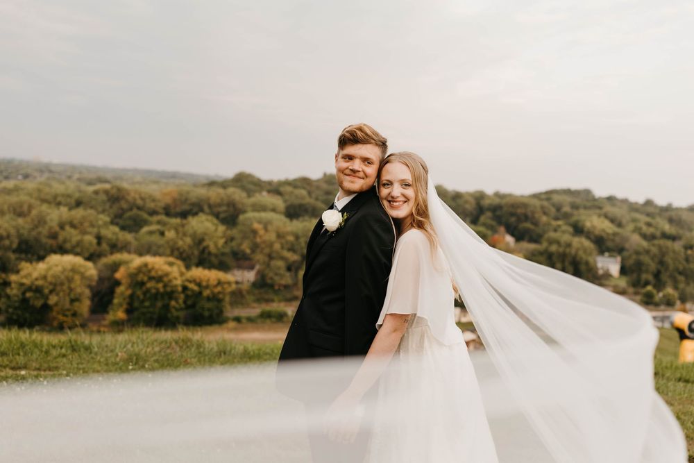 Smiling bride and groom posing back-to-back with flowing veil and scenic countryside backdrop.