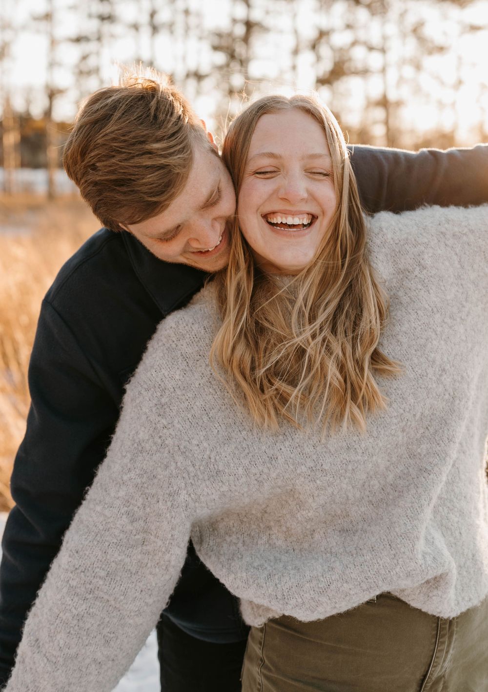 Laughing couple in cozy sweaters embracing outdoors in warm winter sunlight during engagement photos.