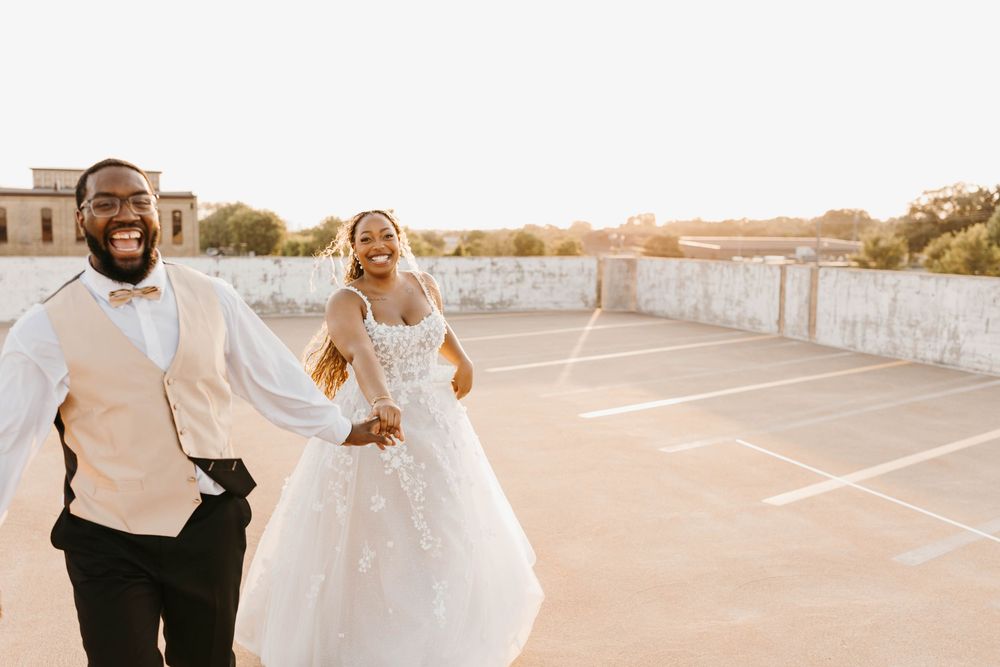 Joyful newlyweds running hand-in-hand on rooftop at sunset, bride in lace gown and groom in vest.