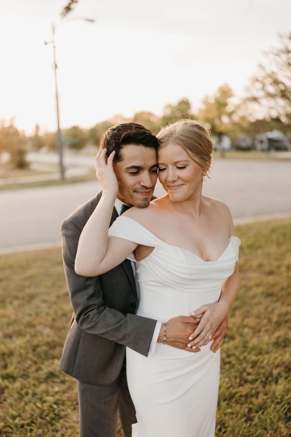 Groom embracing bride from behind at golden hour, bride in off-shoulder white gown on green lawn.