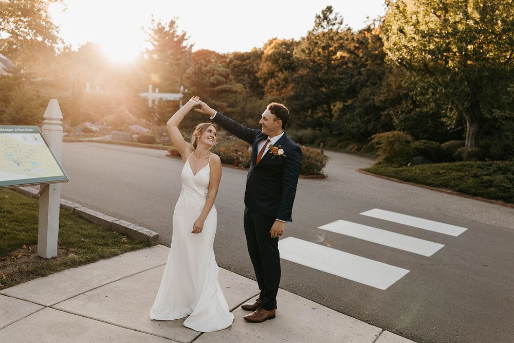 Bride in sleek white gown twirls with groom in navy suit at golden hour on a crosswalk outdoors.