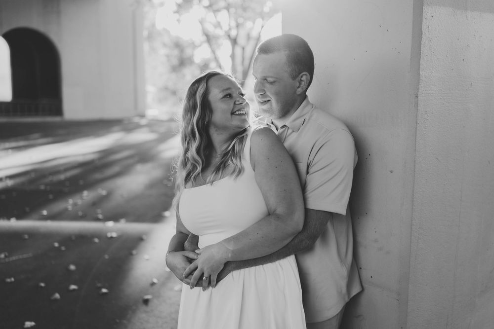 Black and white engagement photo of smiling couple embracing against a wall in soft natural light.
