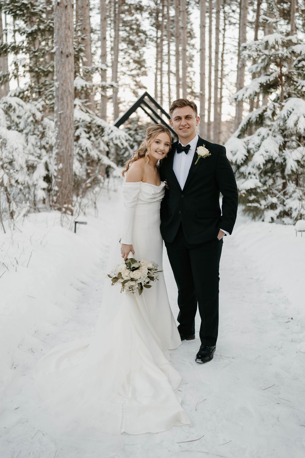 Bride and groom posing in snowy winter forest, bride in off-shoulder gown holding white bouquet.