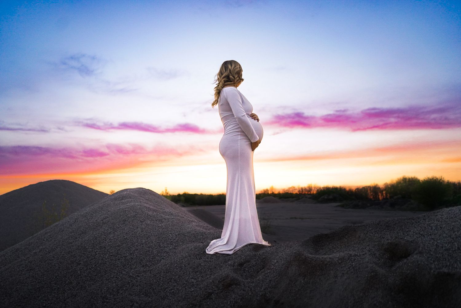 A figure in white stands against colorful sunset sky on desert dunes.